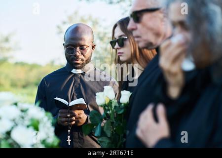 Konzentrieren Sie sich auf einen ernsthaften Priester in schwarzer Kleidung, der beim Beerdigungsgottesdienst in der offenen Heiligen Bibel schaut, während er Verse laut liest und vor dem Sarg betet Stockfoto