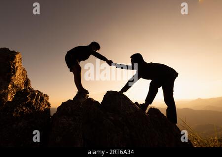 Silhouetten von zwei Menschen, die auf den Berg klettern und helfen. Hilfe- und Assistenzkonzept. Helfende Hand, sportliches Training. Stockfoto