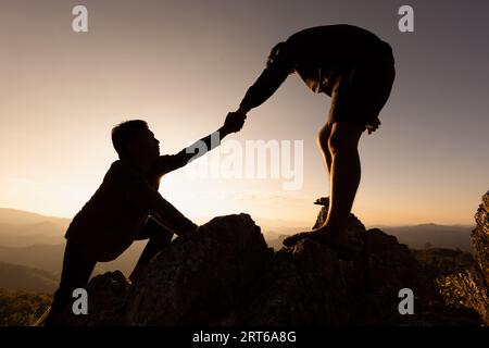 Silhouetten von zwei Menschen, die auf den Berg klettern und helfen. Hilfe- und Assistenzkonzept. Silhouette von Teamwork auf den Bergen Helping Hand, Spor Stockfoto
