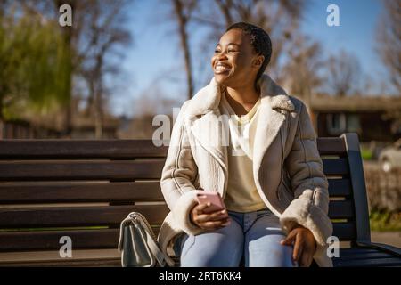 Außenporträt einer glücklichen afroamerikanischen Frau am sonnigen Tag. Sie sitzt auf einer Bank auf der Straße und benutzt ihr Smartphone. Stockfoto