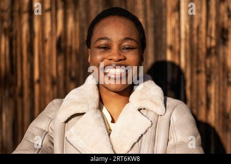 Außenporträt einer glücklichen afroamerikanischen Frau. Sie steht vor einer hölzernen Mauer und lächelt. Stockfoto