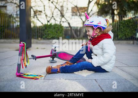 Vorschulmädchen im Einhorn-Helm, das nach dem Sturz auf dem Boden saß, während sie an einem Frühlingstag im Park mit dem Roller fuhr. Outdoor-Sportaktivitäten und Stockfoto