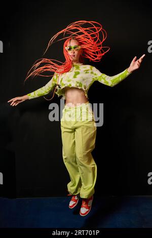 Eine lange, junge, stylische Frau mit hellgrünem Outfit und Sonnenbrille mit bunten Afro-Zöpfe, die über den Boden vor schwarzem Hintergrund springen Stockfoto