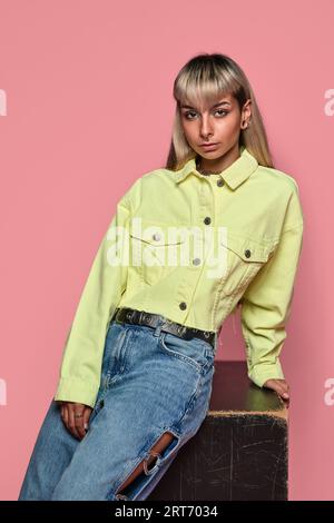 Junger, trendiger weiblicher Hipster mit langen blonden Haaren in einer gelben Jacke und Jeans, die auf einem Würfel auf einem rosa Hintergrund sitzen Stockfoto