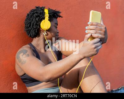 Glückliche ethnische Frau mit geschlossenen Augen in Crop Top sitzt auf orangem Hintergrund und hört Musik in Kopfhörern vom Mobiltelefon im Sonnenlicht Stockfoto