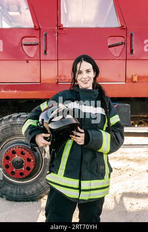 Junge positive Feuerwehrfrau mit schwarzem Schweineschwanzhelm in Uniform gegen rote Feuerwehrauto Stockfoto