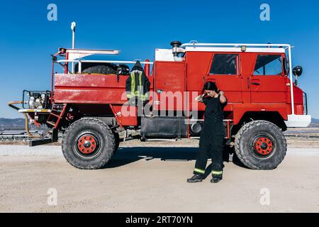 Vollständiger Körper einer weiblichen Feuerwehrfrau, die Arbeitskleidung trägt, die Haare anpasst, während sie sich auf die Arbeit vorbereitet und in der Nähe der roten Feuerwehrmaschine gegen den blauen wolkenlosen Himmel steht Stockfoto