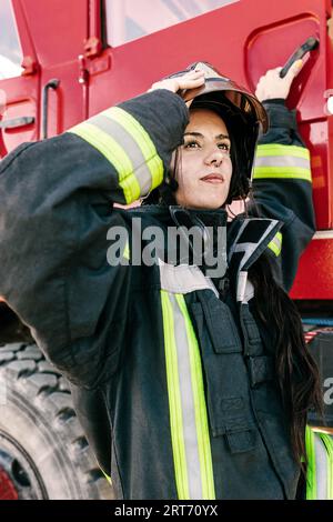 Junge, furchtlose Feuerwehrfrau trägt Arbeitskleidung, die den Schutzhelm anpasst, während sie sich auf die Arbeit vorbereitet, um in die rote Feuerwehrmaschine zu gelangen Stockfoto