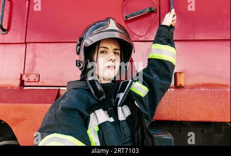 Junge furchtlose Feuerwehrfrau, die Arbeitskleidung und Schutzhelm trägt, um sich auf die Arbeit vorzubereiten und in die rote Feuerwehrmaschine zu gelangen Stockfoto