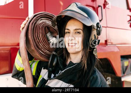 Tapfere Feuerwehrfrau trägt Helm und Uniform und trägt schweren Schlauch auf der Schulter gegen den roten Feuerwehrauto während der Arbeit Stockfoto