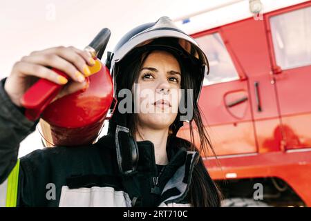 Geringer Winkel einer jungen Feuerwehrfrau, die Schutzkleidung und Helm trägt, während sie während der Arbeit einen schweren Feuerlöscher gegen einen roten Lkw trägt Stockfoto