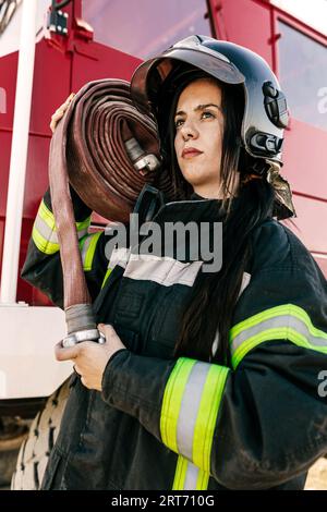 Tapfere Feuerwehrfrau trägt Helm und Uniform und trägt schweren Schlauch auf der Schulter gegen den roten Feuerwehrauto während der Arbeit Stockfoto