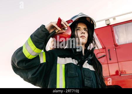 Geringer Winkel einer jungen Feuerwehrfrau, die Schutzkleidung und Helm trägt, während sie während der Arbeit einen schweren Feuerlöscher gegen einen roten Lkw trägt Stockfoto