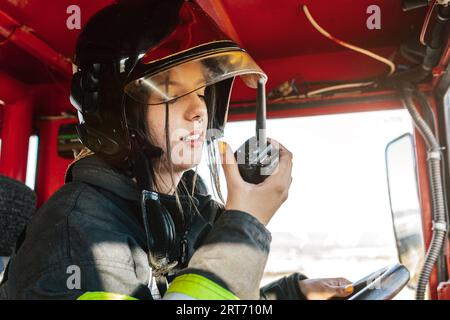 Tapfere Feuerwehrfrau trägt Schutzhelm und Arbeitskleidung, die rote Feuerwehrmaschine fährt, während sie während der gefährlichen Arbeit auf Walkie Talkie spricht Stockfoto