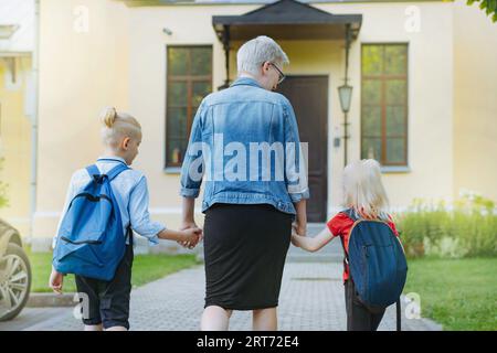 Mutter begleitet Kinder mit Händen zur Schule. Kaukasischer blonder Junge mit im Pferdeschwanz gefesselten Haaren und kleines Mädchen, das mit Mutter zur Schule geht. Bild Stockfoto