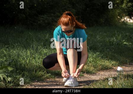 Eine rothaarige Frau, die ihre Schnürsenkel anbindet, bevor sie an einem sonnigen Tag im Park joggt Stockfoto