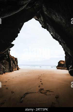 Foto von Fußspuren in der Höhle mit Ozean im Hintergrund bei Sonnenaufgang. Ebbe in Meereshöhle mit goldenem Sand am Strand mit Felsen im Hintergrund. Beau Stockfoto