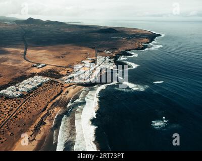 Luftaufnahme des Sonnenuntergangs auf Caleta de Famara auf den Kanarischen Inseln - Lanzarote: Breites Foto von oben mit starken Wellen im blauen Ozean und weißem klarem Himmel. Stockfoto