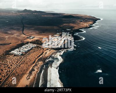 Luftaufnahme des Sonnenuntergangs auf Caleta de Famara auf den Kanarischen Inseln - Lanzarote: Breites Foto von oben mit starken Wellen im blauen Ozean und weißem klarem Himmel. Stockfoto