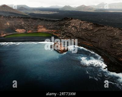 Luftaufnahme des grünen Sees El Golfo auf Lanzarote - Kanarische Inseln. Klippen und Felsen von oben mit blauem Ozean mit Wellen. Dramatisch und stimmungsvoll dunkles Foto Stockfoto