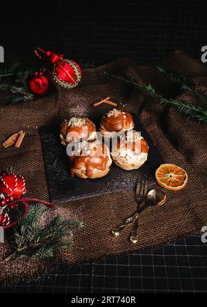 Profiterolen mit weißer Creme und weihnachtsdekoration auf dunklem Hintergrund. Vintage-Foto von leckerem Dessert mit Karamell - rustikaler Stil. Stockfoto