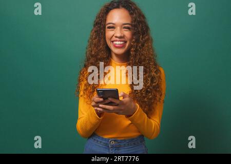Schöne junge Frau, die auf dem Handy SMS schreibt, lächelnd, grüner Studiohintergrund Stockfoto