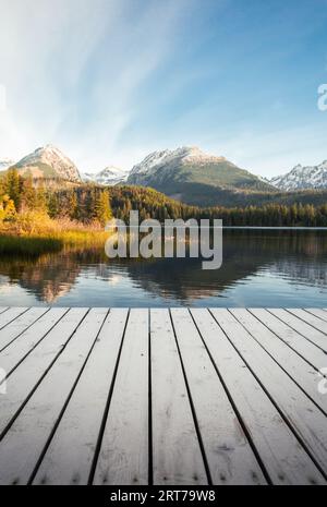 Vertikales Foto - Pier auf dem See in Herbstlandschaft und wunderschöne und verschneite Berge im Hintergrund. Strbske pleso in der Hohen Tatra im Herbst mit Holz Stockfoto