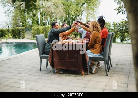 Eine vielfältige Gruppe von Freunden, darunter kaukasier und ein afrikanisches Individuum, toasten mit Weingläsern in einem Gartenhof am Pool. Sie sitzen zusammen Stockfoto