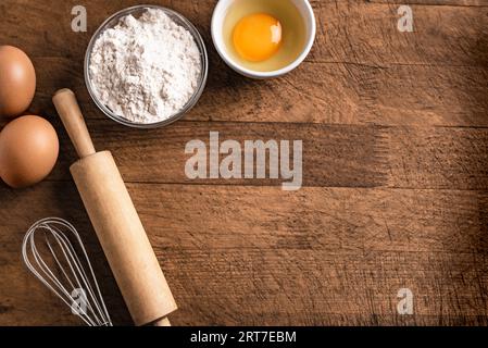 Weizenmehl, Eier, Stecknadel und Schneebesen auf hölzernem Hintergrund, Draufsicht, Kopierraum. Zutaten für Bäckerei, Backen oder Kochkonzept. Stockfoto