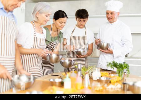 Kochkurs - männlicher Koch in Kochuniform bringt jungen Leuten Kochkurse bei, um Zutaten für Gerichte in der Restaurantküche zuzubereiten und zu mischen Stockfoto