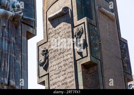Eine georgische Inschrift in Mkhedruli-Schrift füllt eine steinerne Plakette in Form einer Schriftrolle auf einem Pfeiler der Chronik von Georgien (georgisch საქართველოს მატიანე, Sakartvelos Matiane) auf dem Berg Kenisi über dem Tbilisi-See, einem Stausee, im Bezirk Nadzaladevi von Tiflis, Georgien. Kleine Bronzebilder von Engeln flankieren die Plakette. Der Rand der Plakette rollt sich oben über. Links hält eine große Bronzefigur einen Gegenstand in den Händen. Rechts trägt die Pfeilerwand eine Schriftrolle mit einer verwitterten Inschrift. Der Text auf der Plakette gibt Daten vom späten 13. bis zum frühen 15. Jahrhundert an, einschließlich 1346–1360 und  Stockfoto