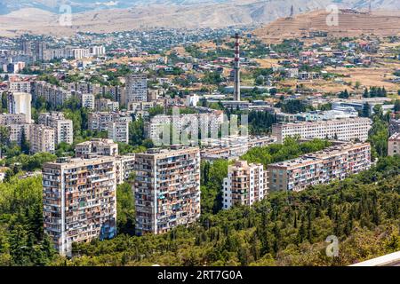 Blick vom Kenischberg auf die Wohnsiedlungen aus der Sowjetzeit in der Metropolregion Tiflis. Die weitläufigen vorgefertigten Siedlungen aus der Sowjetzeit dominieren große Teile der georgischen Hauptstadt, die eine Million Einwohner hat; im Hintergrund ein Kamin und die Hügelketten am Stadtrand, umrahmt von den Vorgebirgen des Kaukasus. Die Chronik von Georgien ist ein von weitem sichtbares Monument auf dem Berg Keeni bei Tiflis, der Hauptstadt Georgiens. Sie wurde 1985 vom Bildhauer Zurab Tsereteli geschaffen. Sanzona, Bezirk Nadzaladevi, Tiflis, 0191, Georgien Stockfoto