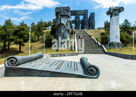 Treppe zum Monument „Chronik von Georgien“ auf dem Mount Kenisi bei Tiflis, Georgien. Im Vordergrund befindet sich eine sitzende Bronzesuppe mit einer georgischen Inschrift; links eine gefallene Säule, die Teil des unvollendeten Komplexes ist. Die Arbeiten an dem Denkmal durch den Bildhauer Surab Zereteli (Zurab Tsereteli) begannen 1985; Besucher sind auf der offenen Treppe zu sehen. Utsnob Gmirta Street, Sanzona, Bezirk Nadzaladevi, Tiflis, 0191, Georgien Stockfoto