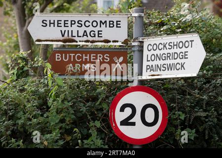 Ländliche Wegweiser zu einem lokalen Pub und nahe gelegenen Dörfern sowie eine Geschwindigkeitsbegrenzung von 20 km/h im Cotswolds-Dorf Sheepscombe, am 8. September 2023, in der Nähe von Painswick, England. Seit Anfang des 17. Jahrhunderts war Sheepscombe wie viele der Cotswold-Städte in der Gegend und dem benachbarten Painswick an der Tuchherstellung beteiligt. Stockfoto