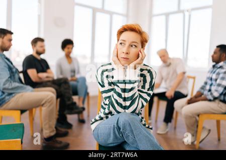 Porträt einer depressiven jungen Frau, die Ohren mit Händen bedeckt, die in der gruppenpsychologischen Beratung im Kreis auf die Kamera schauen. Stockfoto