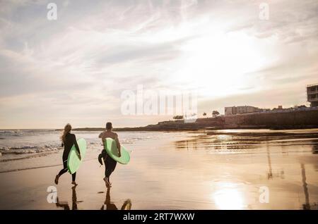 Surferpaar, das bei Sonnenuntergang mit Surfbrettern am Strand entlang läuft Stockfoto