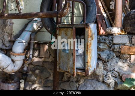 Eine offene, rostige Metallbox ist an einer Wand befestigt. Es ist ein Messgerät in der Box. Es gibt rostige Rohre und alte Reifen in der Gegend. Die Lage ist Chugureti, Tiflis, Georgien Stockfoto