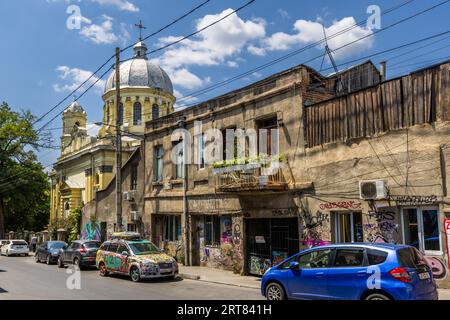 Verwitterte alte Gebäude, die mit Graffiti bedeckt sind, säumen eine Straße im Chugureti-Bezirk von Tiflis, Georgien. Hinter ihnen erhebt sich eine gelb gestrichene Kirche mit einer silbernen Kuppel und einem Glockenturm (möglicherweise die russisch-orthodoxe Alexander-Nevski-Kirche). Auf einem Balkon wachsen Pflanzen in Kästen. Ein Auto, das komplett mit Graffiti bemalt und mit einem Dachkoffer ausgestattet ist, steht am Bordstein. Ein kleines blaues Auto steht rechts. Stromleitungen spannen sich über die Straße. Die Alexander-Nevski-Kirche wurde 1864 mit Spenden der Gläubigen erbaut, und der Glockenturm wurde 1886 hinzugefügt. Stockfoto