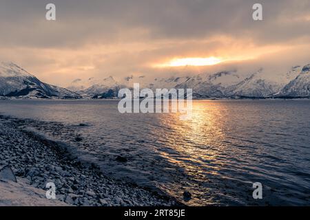 Malerischer Blick über die Morgensonne, die durch dichte Wolken hinter schneebedeckten Bergen in der Nähe des Sees auf dem Archipel der Lofoten in Norwegen bricht Stockfoto