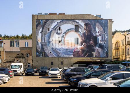 Ein Wandgemälde bedeckt die Seite eines Gebäudes in Tiflis, Georgien. Das Gemälde zeigt architektonische Elemente, Wolken und Menschen. Autos parken vor dem Gebäude Stockfoto