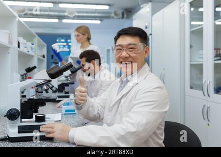 Porträt eines lächelnden und jungen asiatischen Arztes, Wissenschaftler, der hinter einem Mikroskop sitzt und mit dem Superfinger auf die Kamera zeigt, Kollegen arbeiten im Labor dahinter. Stockfoto