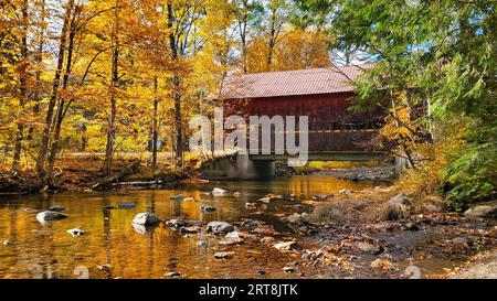 Rote, hölzerne, überdachte Brücke mit schönen Herbstfarben und Flußreflexen, Stowe, Vermont, USA Stockfoto