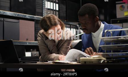 Verschiedene Ermittler führen vertrauliche Untersuchungen durch, analysieren Beweise und recherchieren Aufzeichnungen. Im Büro arbeiten kaukasische und afroamerikanische Detektive zusammen, um Verbrechen zu lösen. Stockfoto