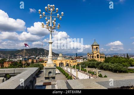 Von der Terrasse der Sameba-Kathedrale (Heilige Dreifaltigkeitskathedrale, Tsminda Sameba) erstreckt sich der Blick über das Kirchenareal und Tiflis, Georgien. Im Vordergrund steht ein hoher Kandelaber mit Globuslampen. Der freistehende Glockenturm erhebt sich rechts, das Torgebäude mit einer kleinen Kuppelkirche geradeaus. Die georgische Flagge mit ihren fünf roten Kreuzen weht links. Am Horizont trägt ein Berg einen Fernsehurm. Die Kathedrale wurde von 1995 bis 2004 auf dem Elia-Hügel im Bezirk Avlabari erbaut. Sie ist die Hauptkirche der georgischen orthodoxen Kirche. Mit 86,1 Metern bis zur Spitze ihres c Stockfoto