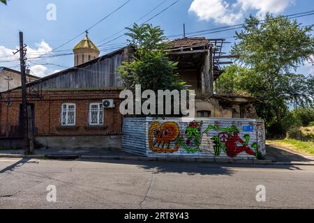 Ein verfallenes Haus mit eingestürztem Obergeschoss steht in einer Straße im Bezirk Chugureti in Tiflis, Georgien. Ein Wellblechzaun, der mit bunten Graffiti-Figuren bemalt ist, schützt die Ruine. Die goldene Kuppel der Sameba-Kathedrale (Tsminda Sameba) erhebt sich hinter dem Dach. Die Kathedrale wurde von 1995 bis 2004 auf dem nahe gelegenen Elia-Hügel im Bezirk Avlabari erbaut. Stockfoto