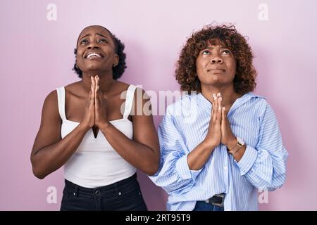 Zwei afrikanische Frauen, die auf rosafarbenem Hintergrund stehen, betteln und beten mit den Händen zusammen mit dem Ausdruck der Hoffnung im Gesicht, sehr emotional und besorgt. Bettler Stockfoto