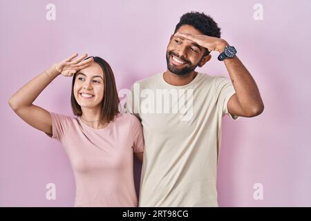 Ein junges hispanisches Paar auf rosafarbenem Hintergrund, sehr glücklich und lächelnd, mit der Hand über dem Kopf weit weg schauend. Konzept wird gesucht. Stockfoto