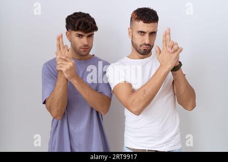 Homosexuelles schwules Paar, das auf weißem Hintergrund steht und eine symbolische Waffe mit Handgeste hält, tötende Schusswaffen spielt, wütendes Gesicht Stockfoto