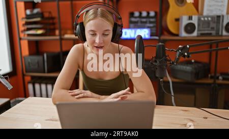 Junge blonde Radioreporterin, die im Podcast-Studio mit Kopfhörern und Laptop spricht Stockfoto