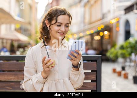 Lächelnde junge Frau, die auf dem Smartphone Text eingibt, Nachrichten beantwortet und online chattet. Sie sieht aus wie eine Social-Media-App auf dem Mobilbildschirm. Ein Touristenmädchen sitzt auf einer Bank auf der Straße der Stadt und trinkt kalten Kaffee mit Eis Stockfoto